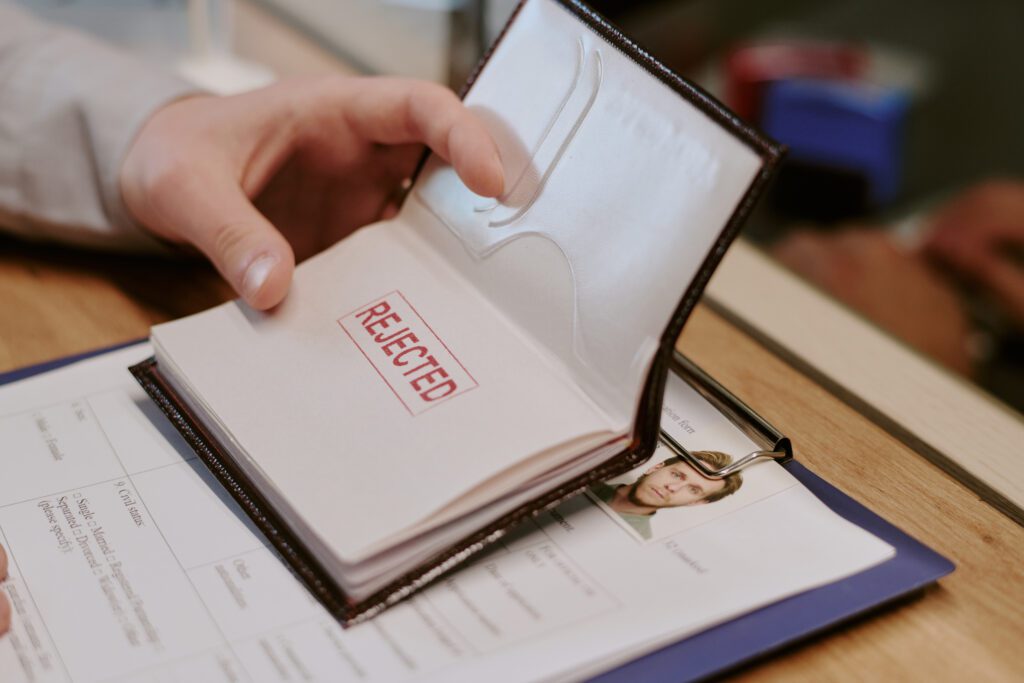 Hand opening passport displaying large red rejected stamp on page accompanied by some personal documents and clipboard with papers on table
