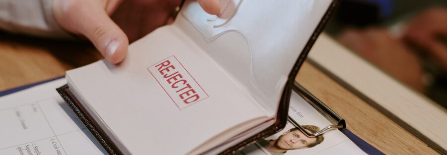 Hand opening passport displaying large red rejected stamp on page accompanied by some personal documents and clipboard with papers on table